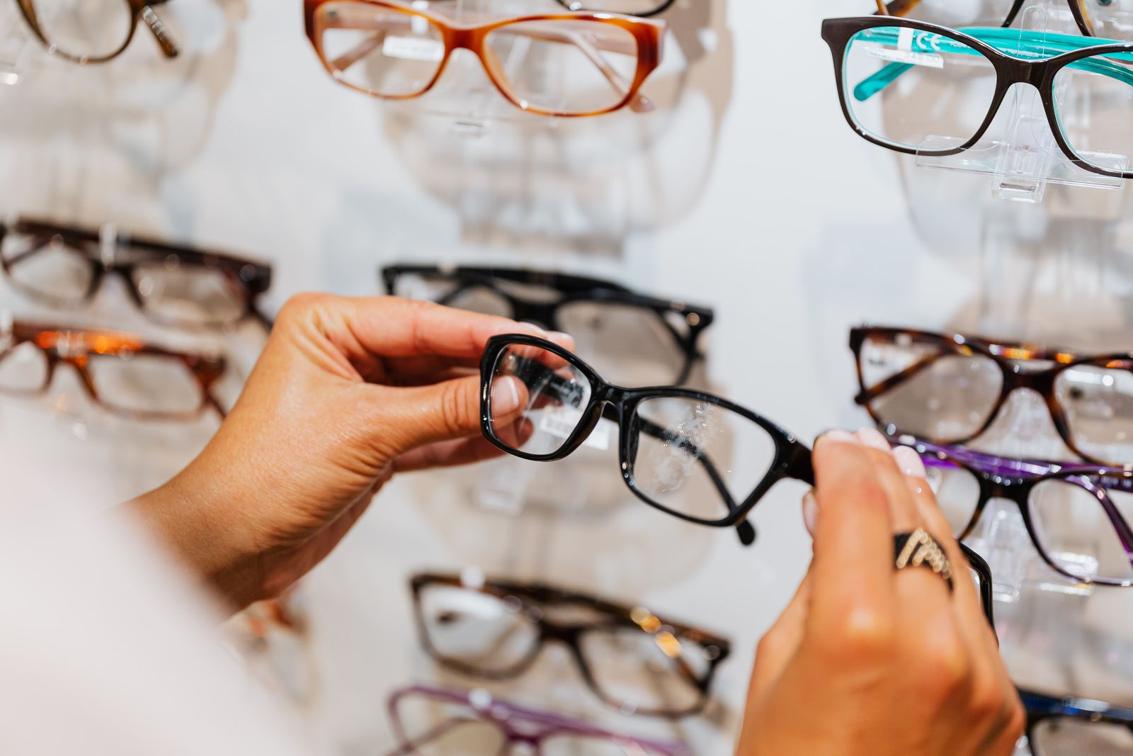 Hands holding a pair of black eyeglass frames up against a display wall